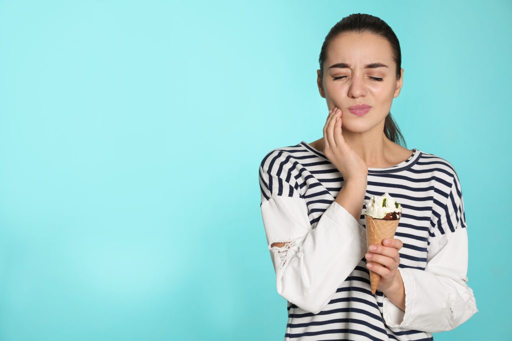 What causes sensitive teeth? Photo of a young woman holding an ice cream in one hand and her jaw in pain with the other hand. 